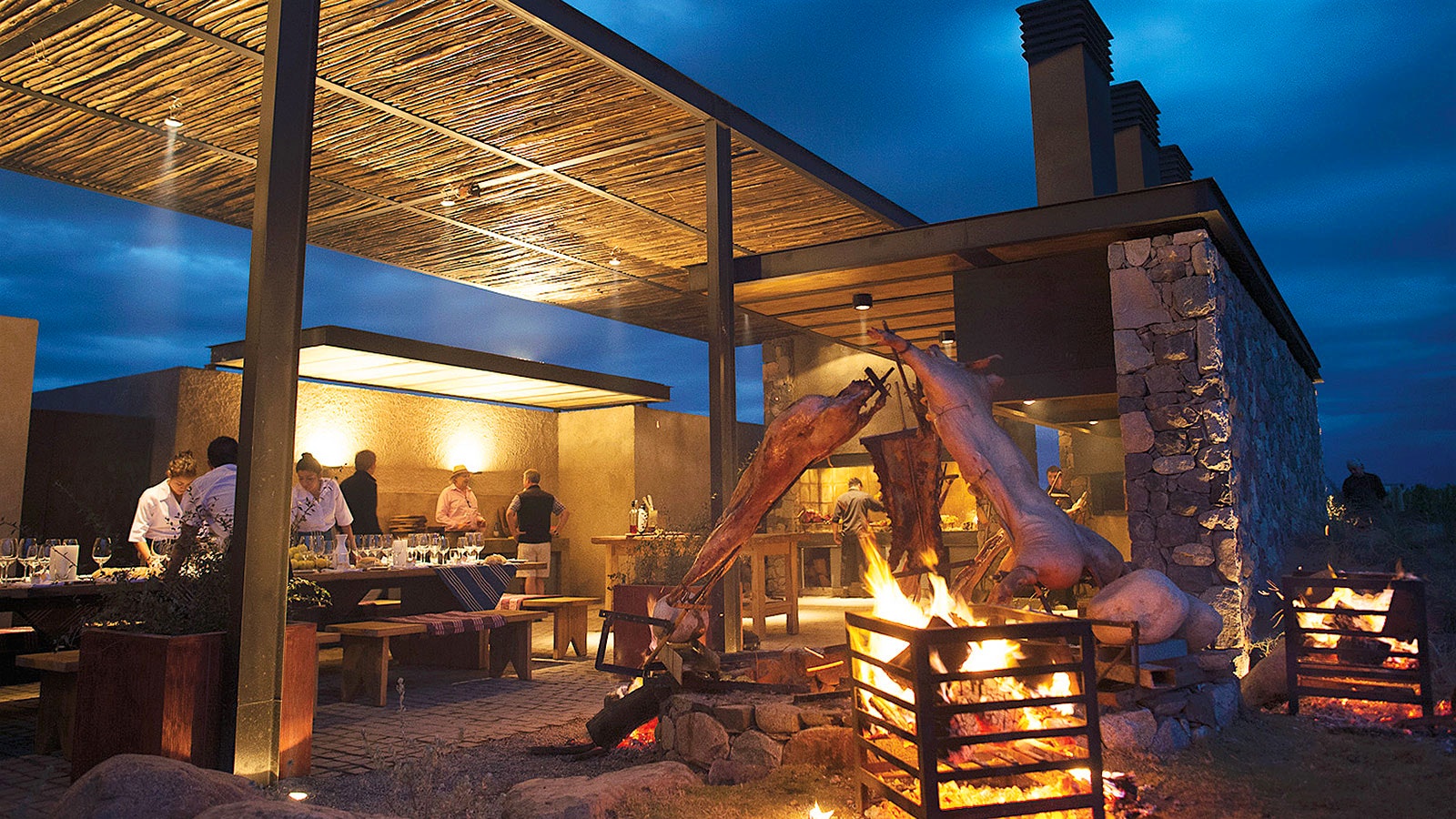 People dining outside at Siete Fuegos beneath a wooden roof, near pigs roasting on an open flame